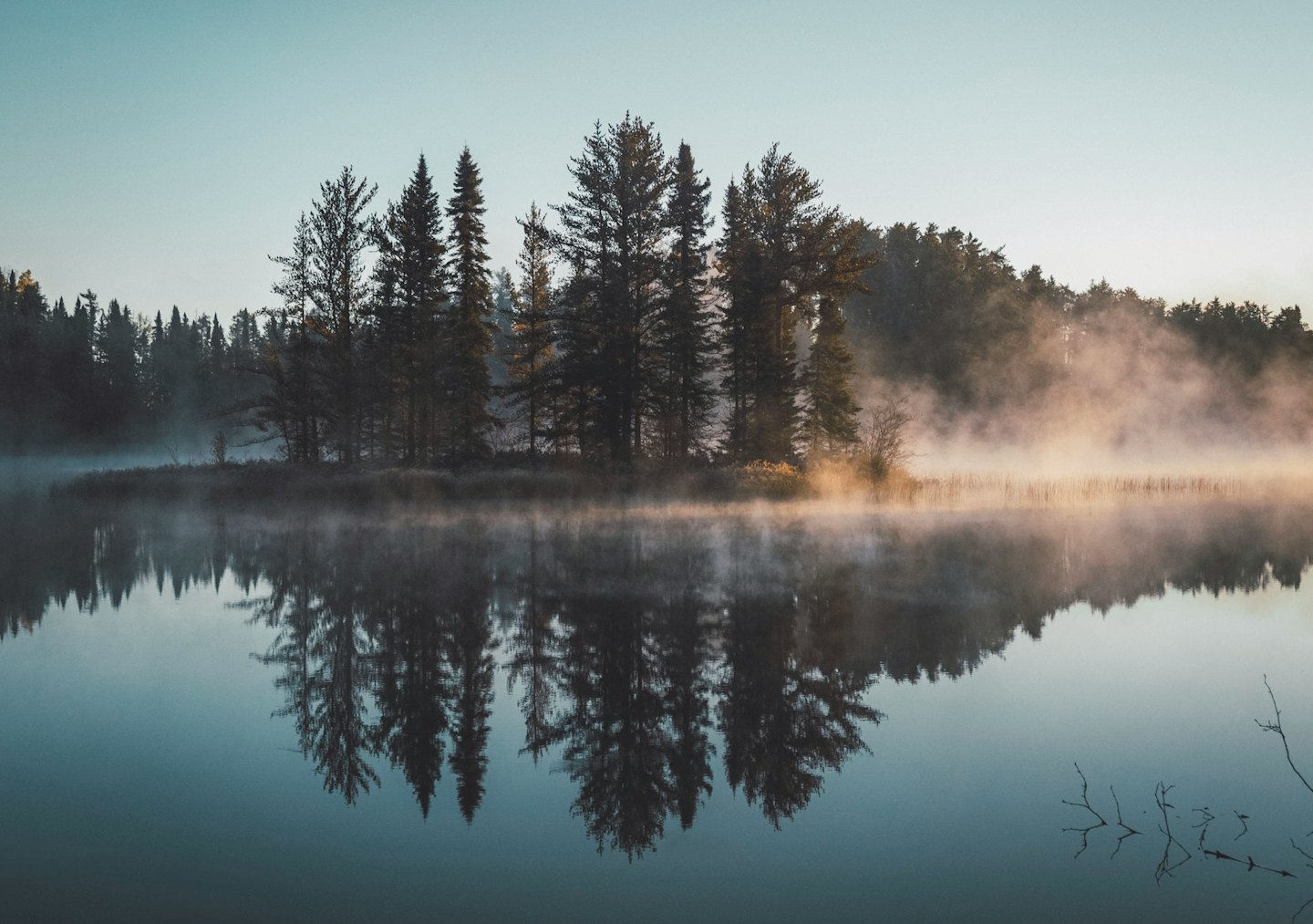 Misty lake at dawn with pine trees reflected in water.